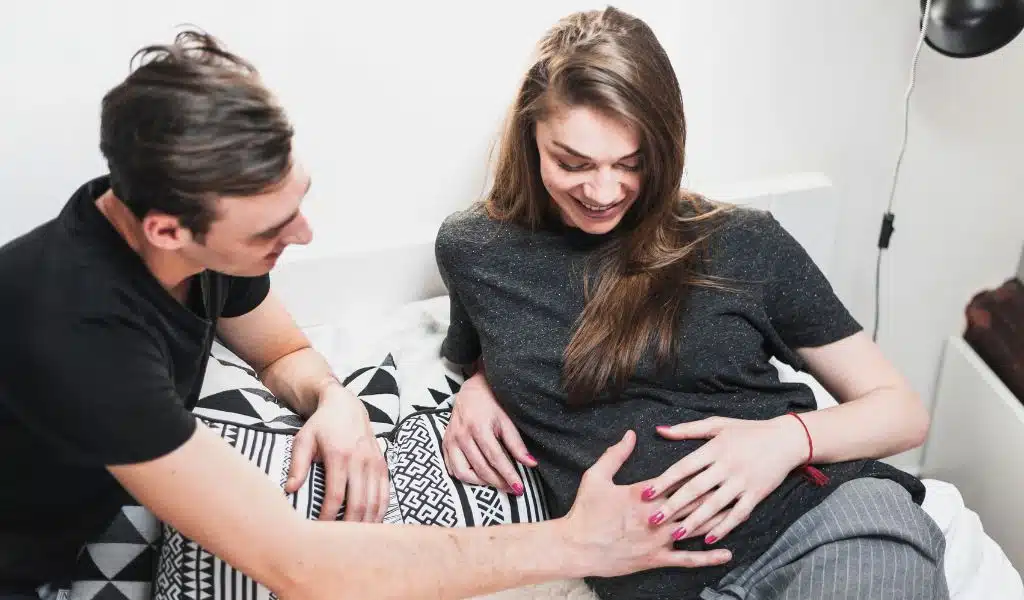 Jeune coupe allongée sur un lit, le jeune papa souriant pose sa main sur le ventre de la jeune femme en teeshirt